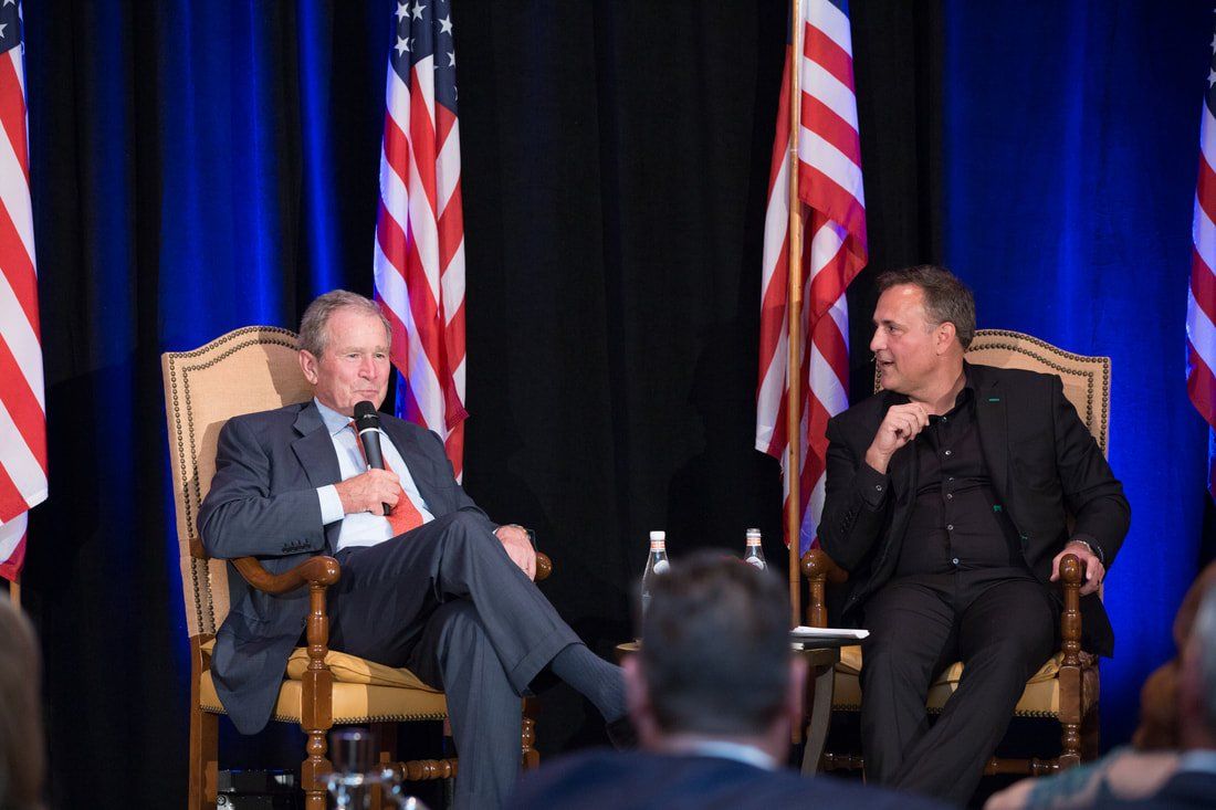 Two men are sitting in chairs on a stage talking to each other.