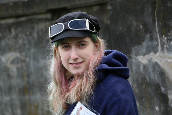 A young woman wearing a hat and sunglasses is standing in front of a wall.