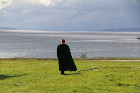 A man in a black cape is standing in a grassy field near the ocean.