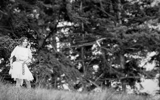 A woman in a white dress is standing in a field surrounded by trees.