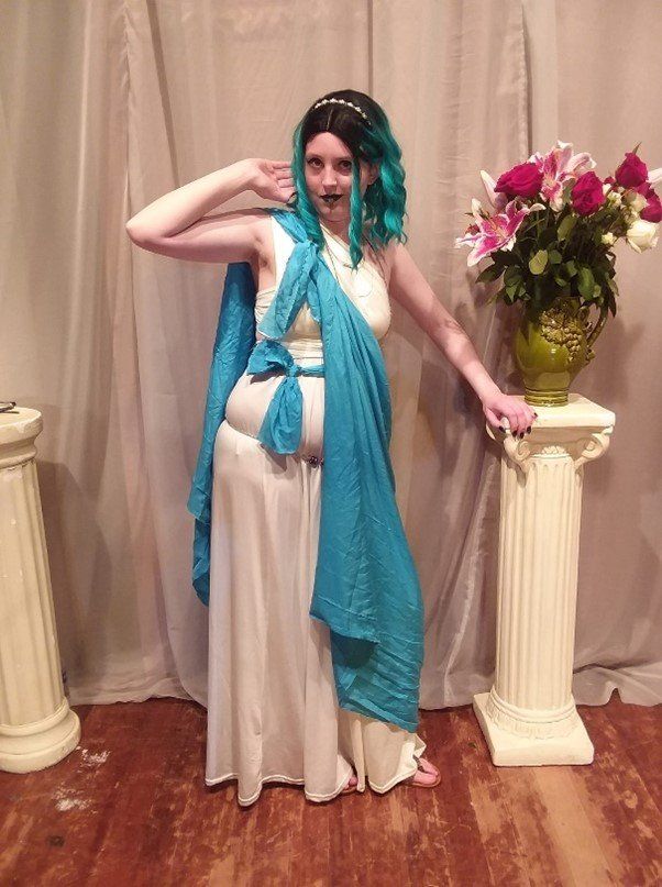 A woman in a costume is standing next to a vase of flowers, Kel representing Aglaia.