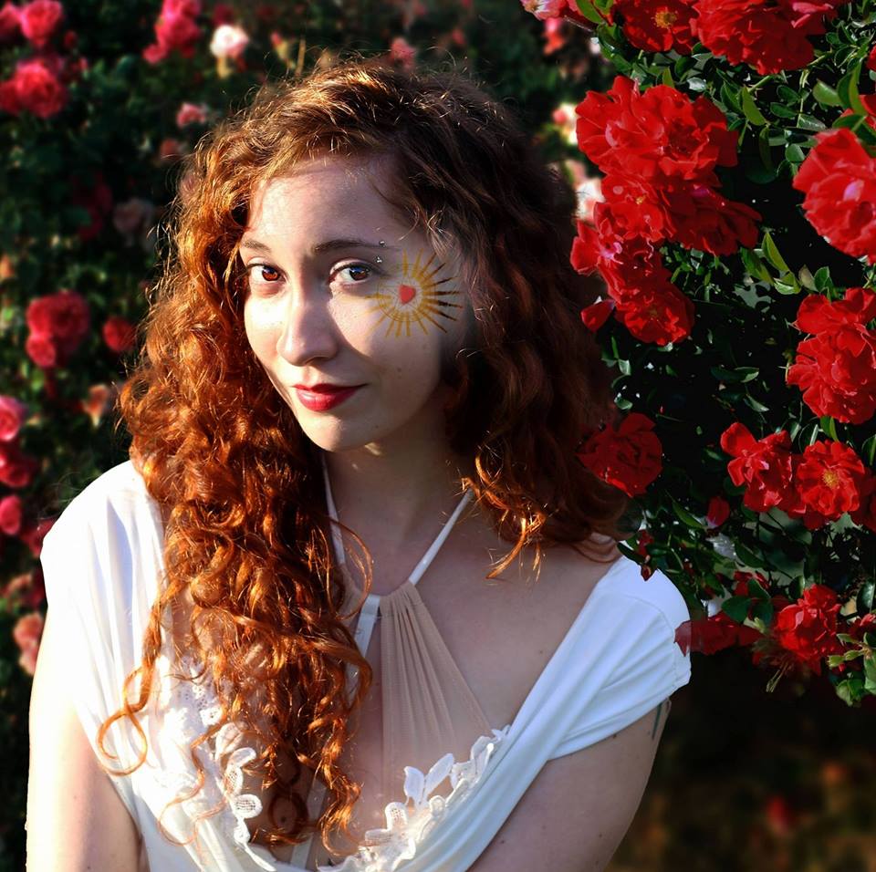 A woman with red hair has a sun painted on her face, Representing Aphrodite.