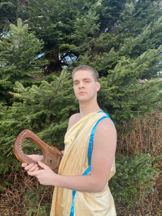 A man in a toga is holding a harp in front of a tree, representing Apollo.