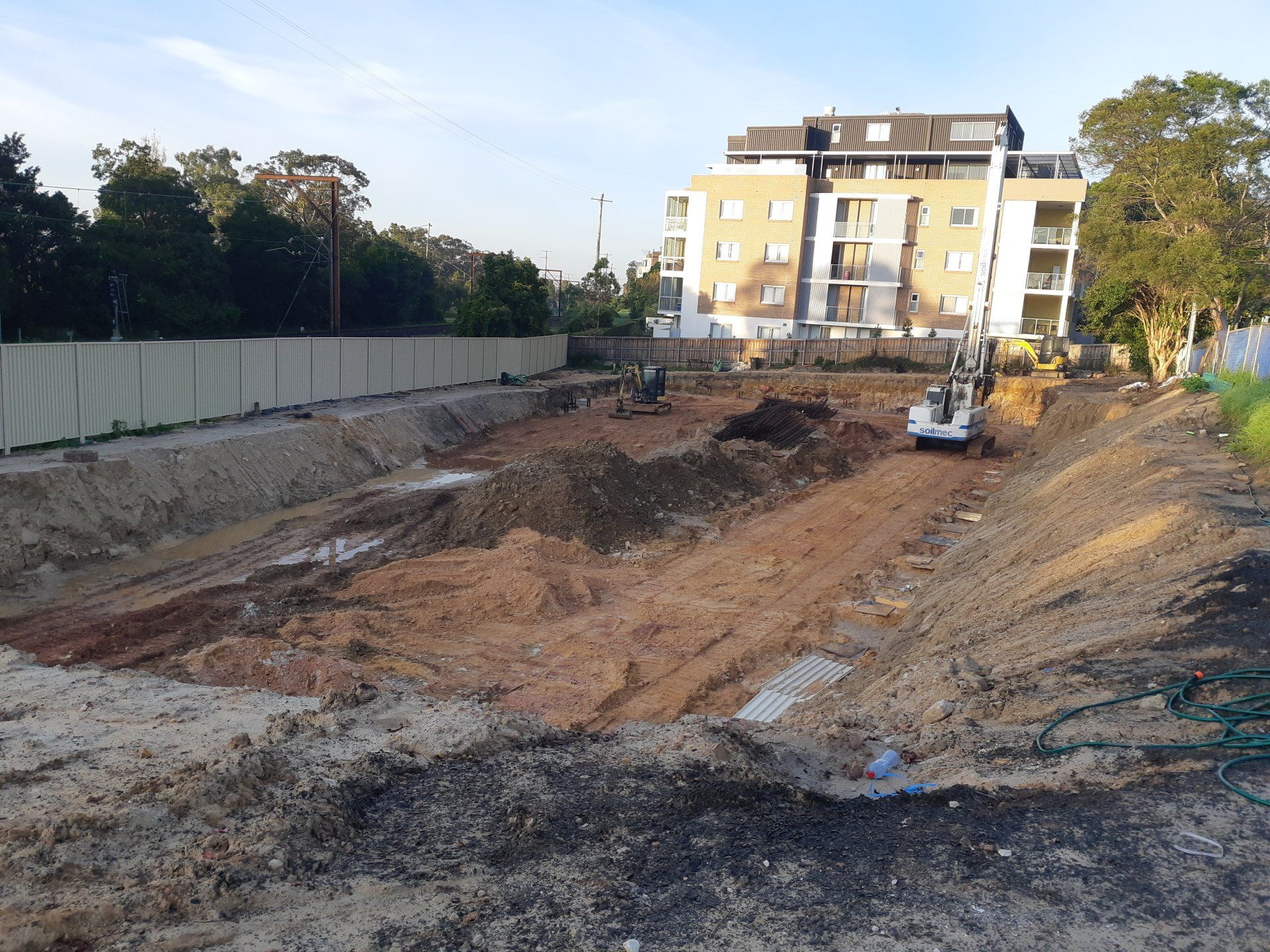Construction site with excavated dirt, heavy machinery, and a yellow building in the background