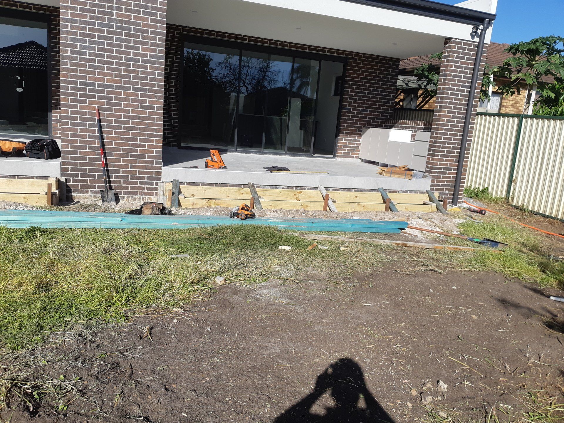 Backyard patio under construction with brick house, concrete slab, dirt foreground, and stacked blocks