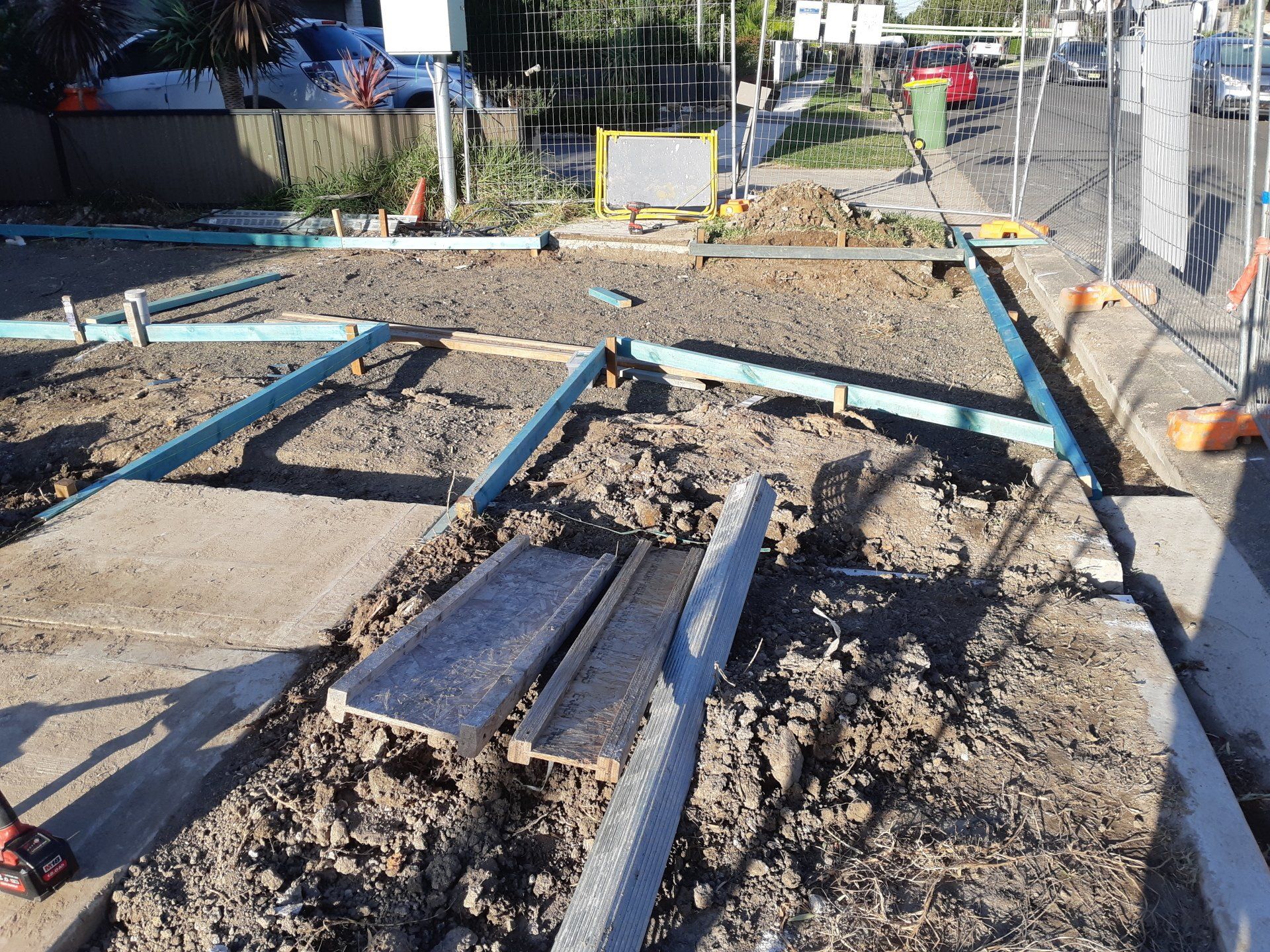 Construction site with blue pipes and wooden planks laid over dirt beside a fence and parked cars