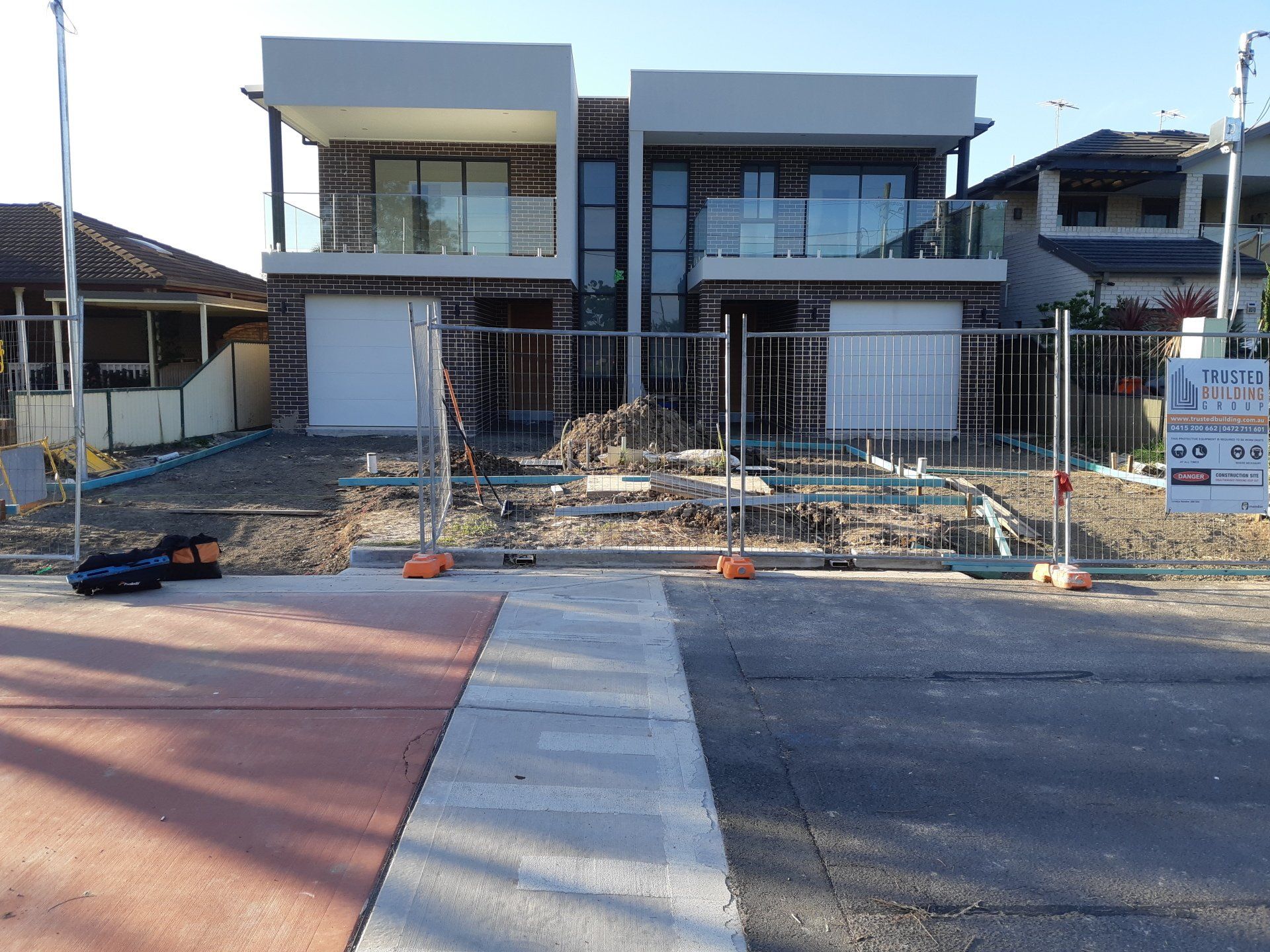 Two-story modern house under construction behind a temporary fence, with paved walkway and unfinished front yard.