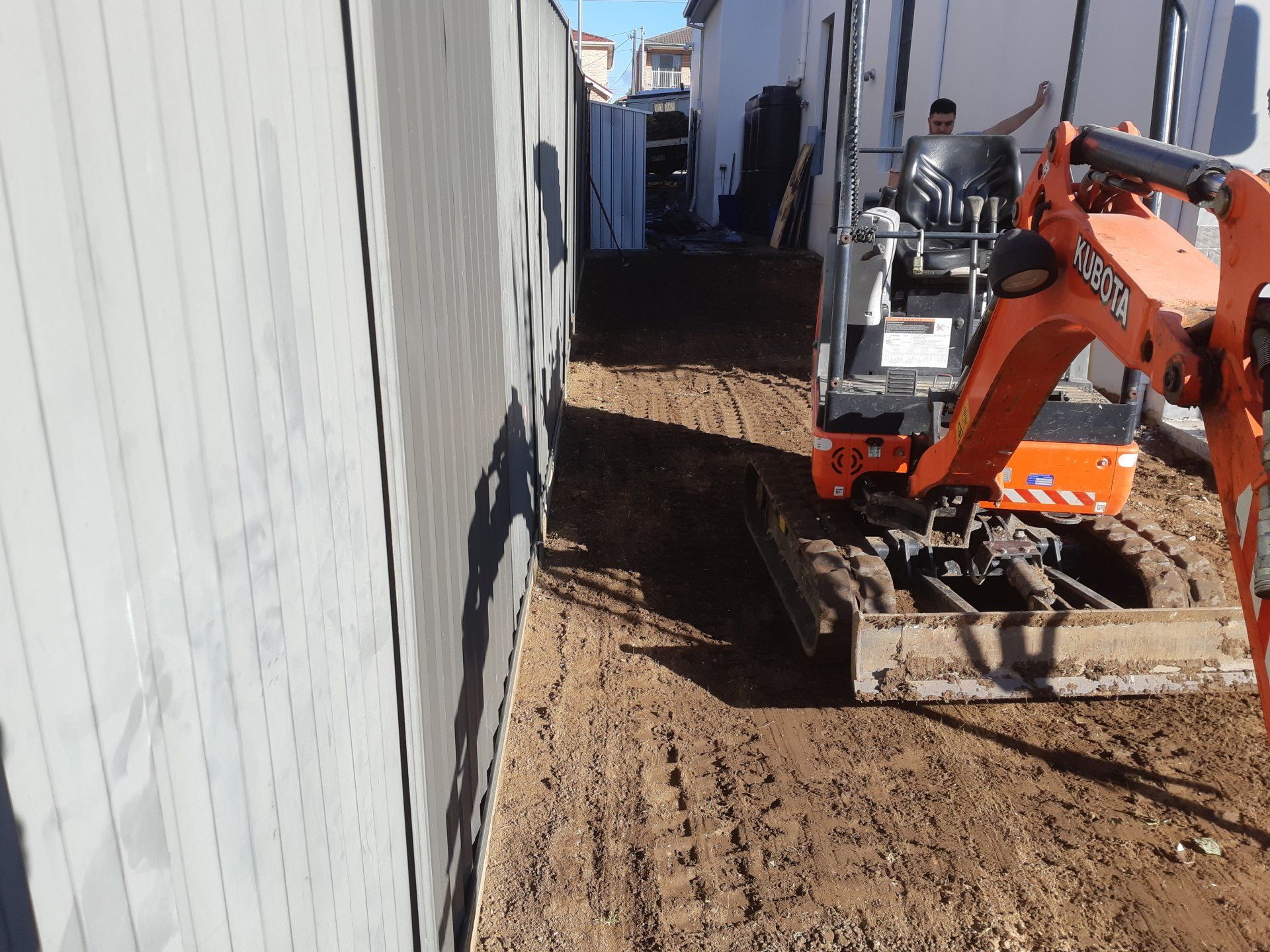 Orange mini excavator digging soil in a narrow alley beside a white building