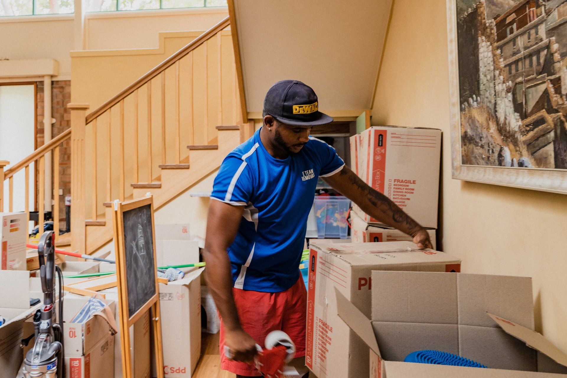 Man in blue shirt and red shorts sorting items in a room with boxes and a staircase