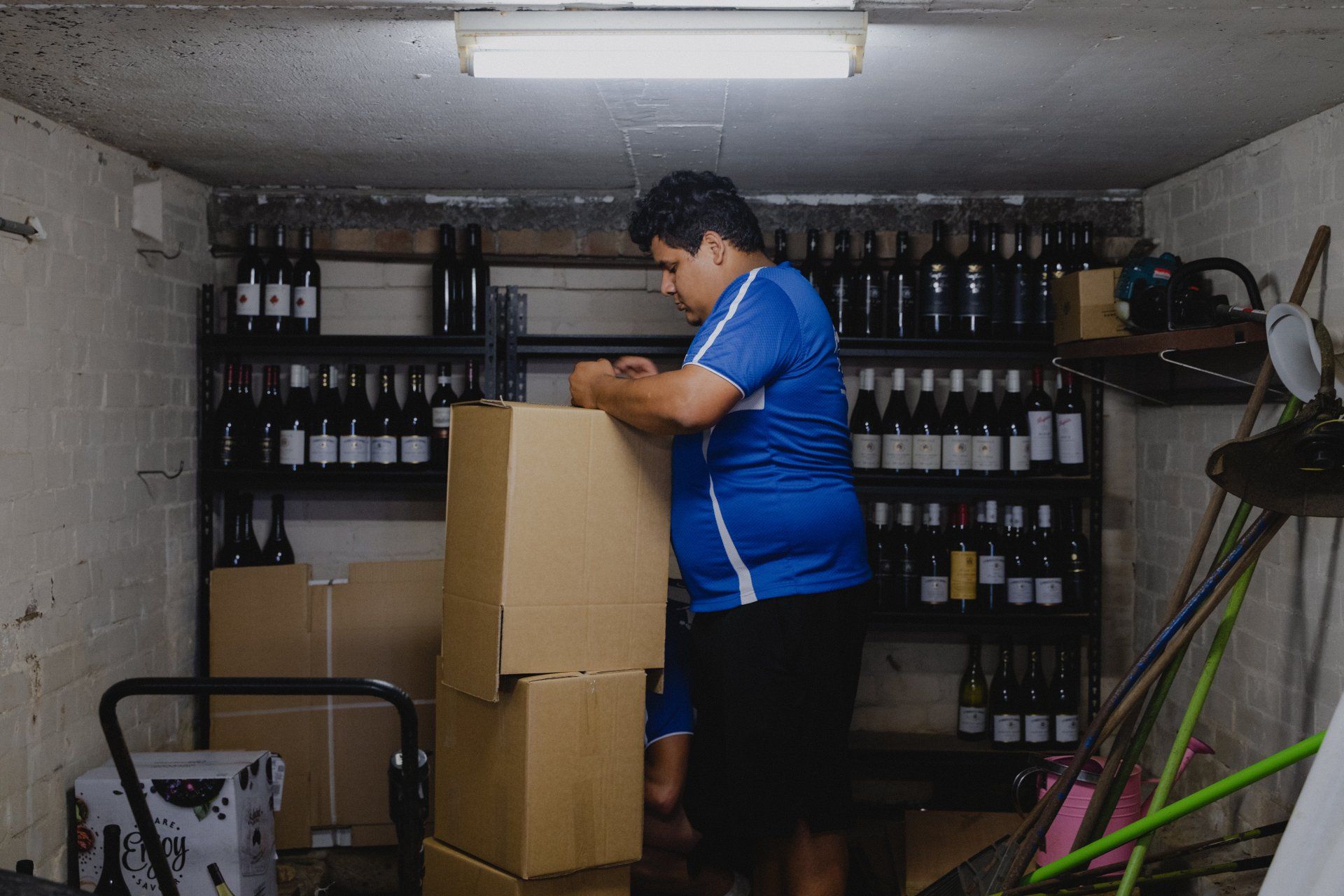 Person packing a cardboard box in a small storage room lined with bottles.