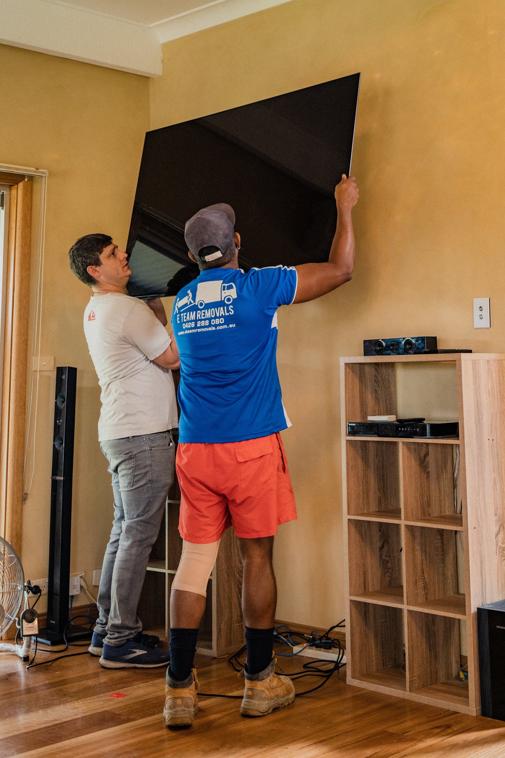 Two people adjusting a large wall-mounted TV in a living room, with cables and a shelf nearby