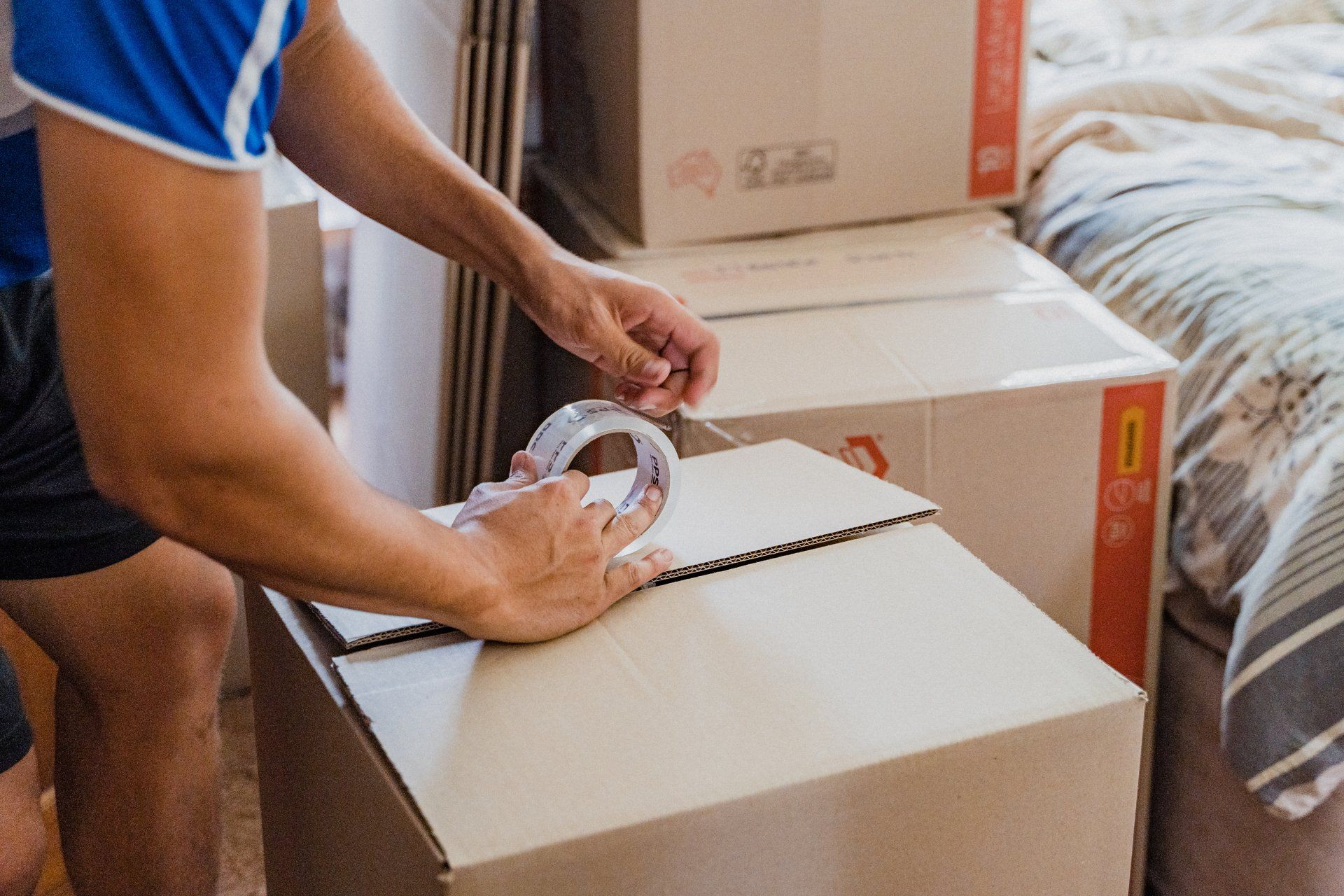 Person sealing a cardboard box with clear packing tape beside stacked moving boxes and a bed