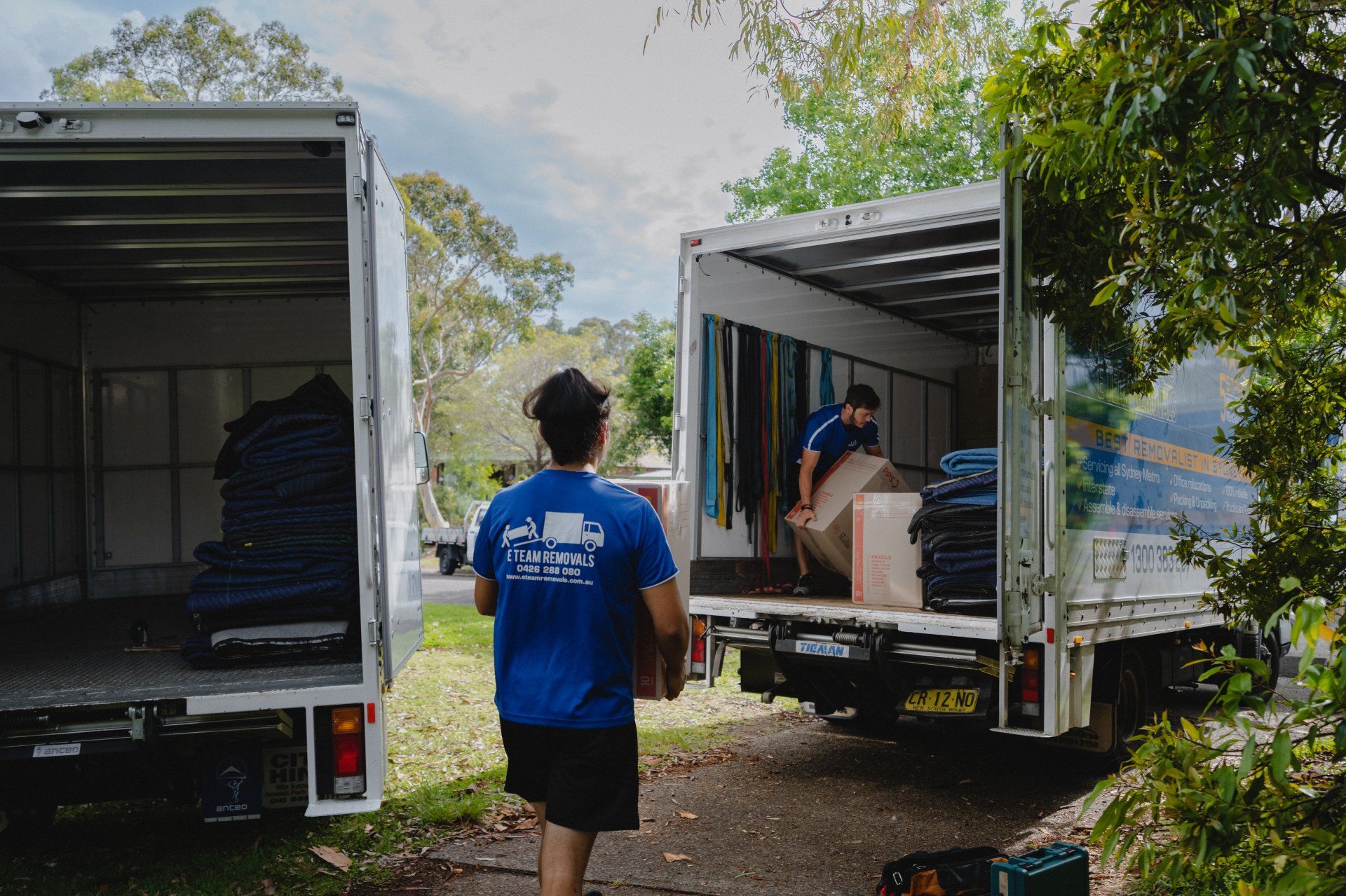 Two moving trucks open in a yard, with a person in a blue shirt carrying items between them.