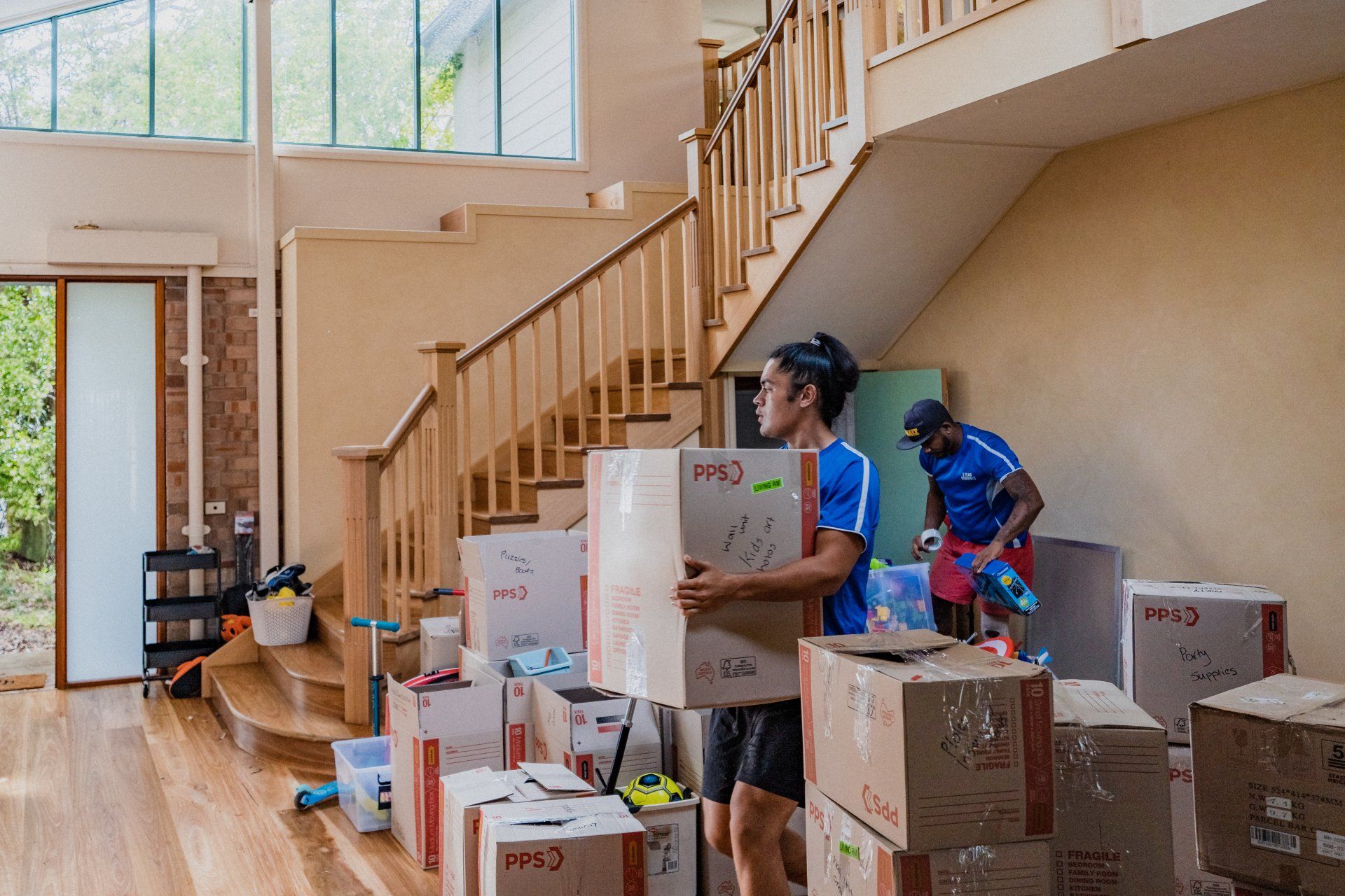 Two people stacking moving boxes in a bright home with a wooden staircase and large windows