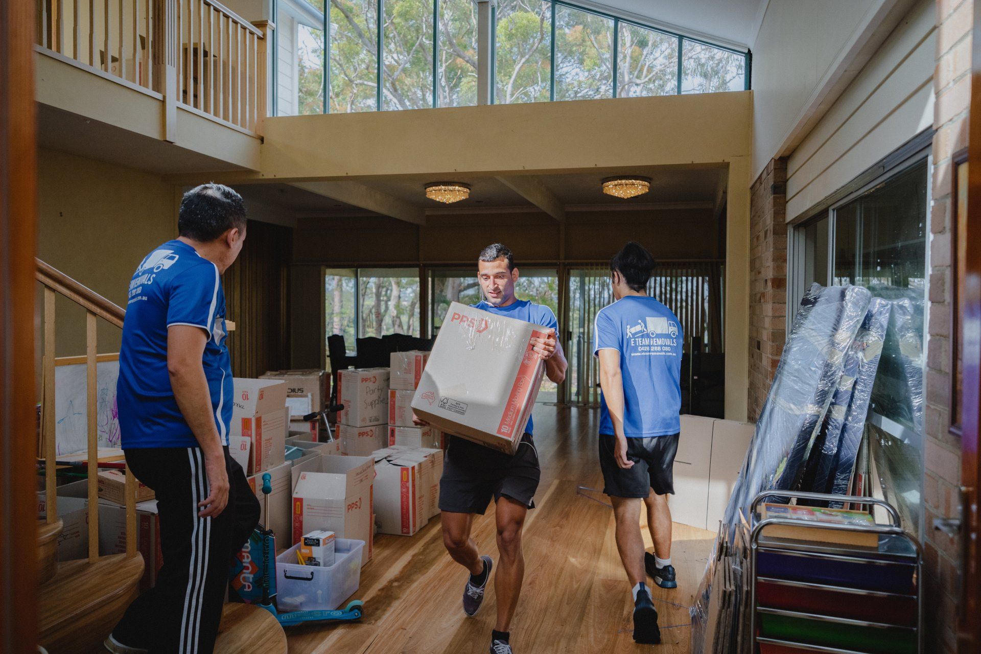 Three people carry moving boxes through a bright hallway with tall windows.