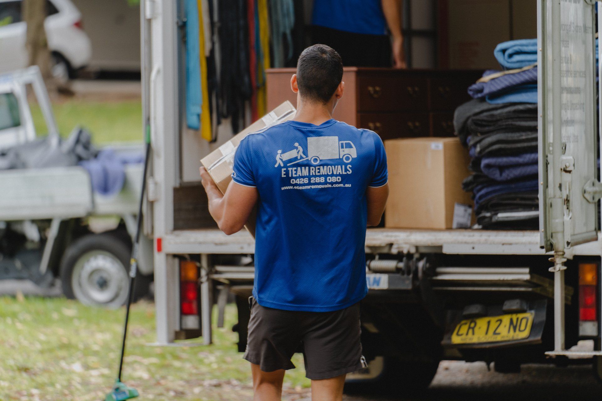 Person in blue shirt loads a box into a moving truck, with stacked blankets and a trailer nearby.