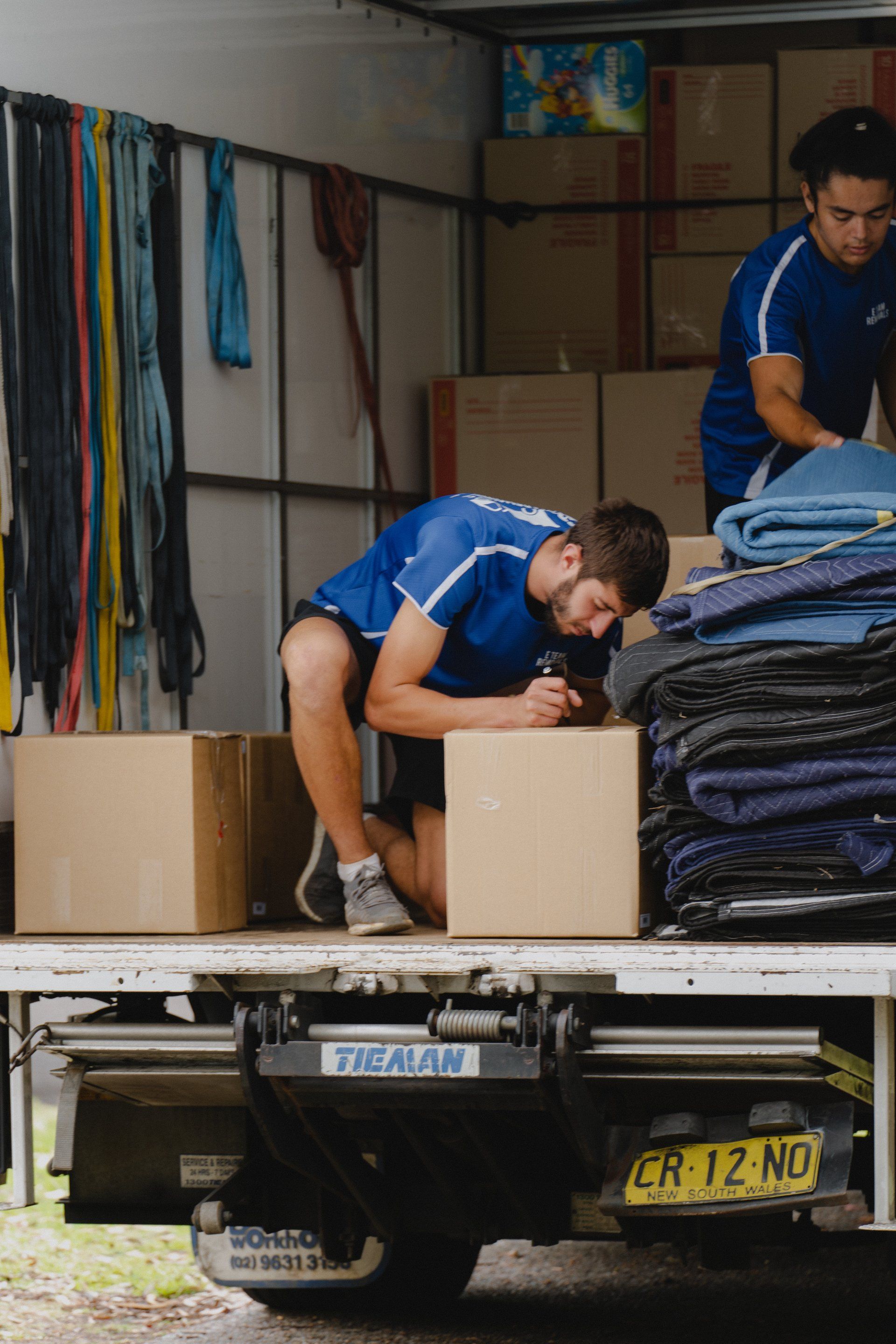 Two men loading stacked blue blankets into a truck from the back of a moving van.