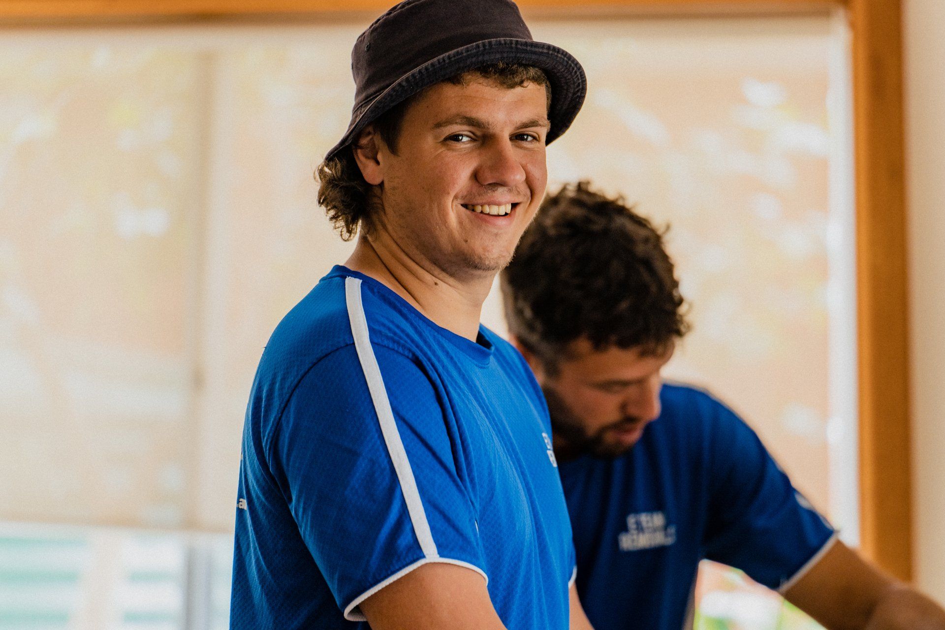 Smiling man in a blue shirt and black hat standing indoors, with another man working in the background