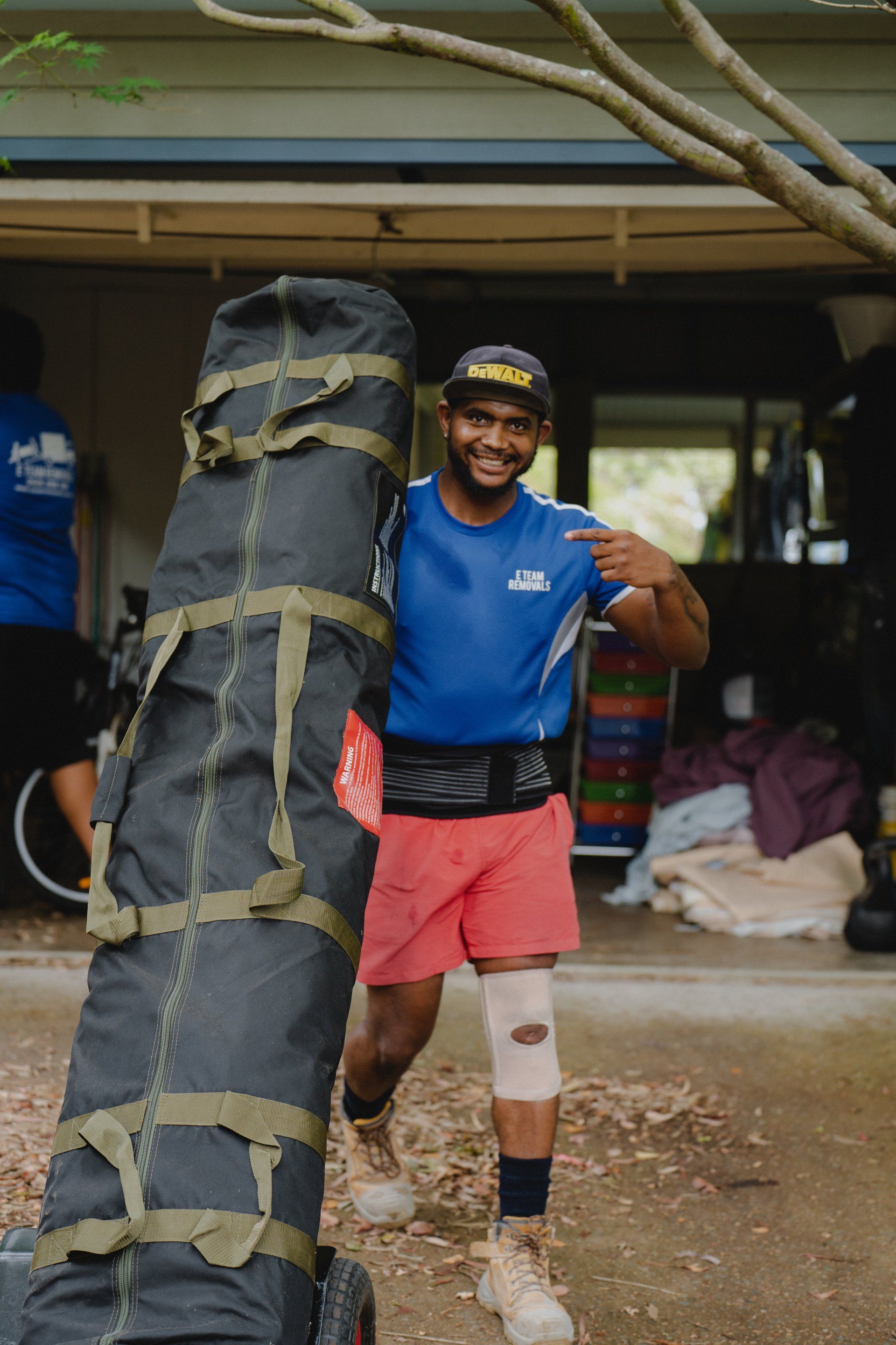 Man in blue shirt and pink shorts carrying a padded training bag outdoors, smiling and posing.