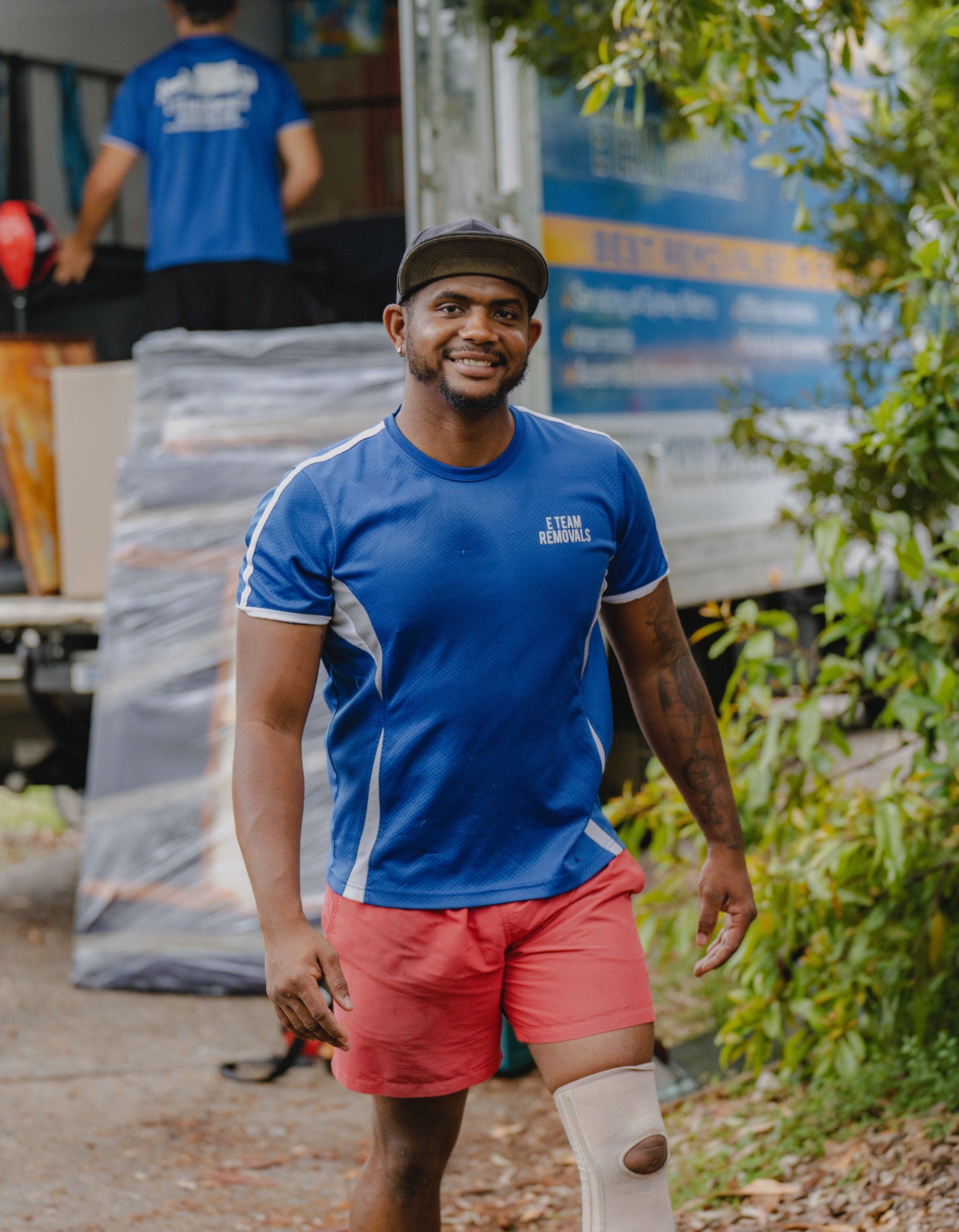 Smiling man in blue shirt and red shorts walking outdoors with a bandaged knee, near stacked boxes.