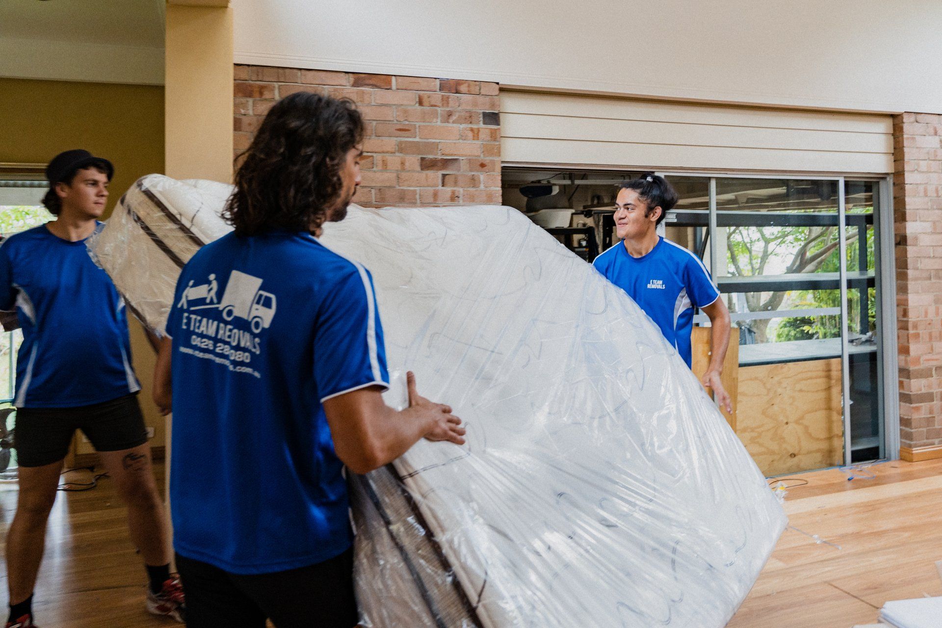 Three movers in blue shirts carry a wrapped mattress through a home interior.