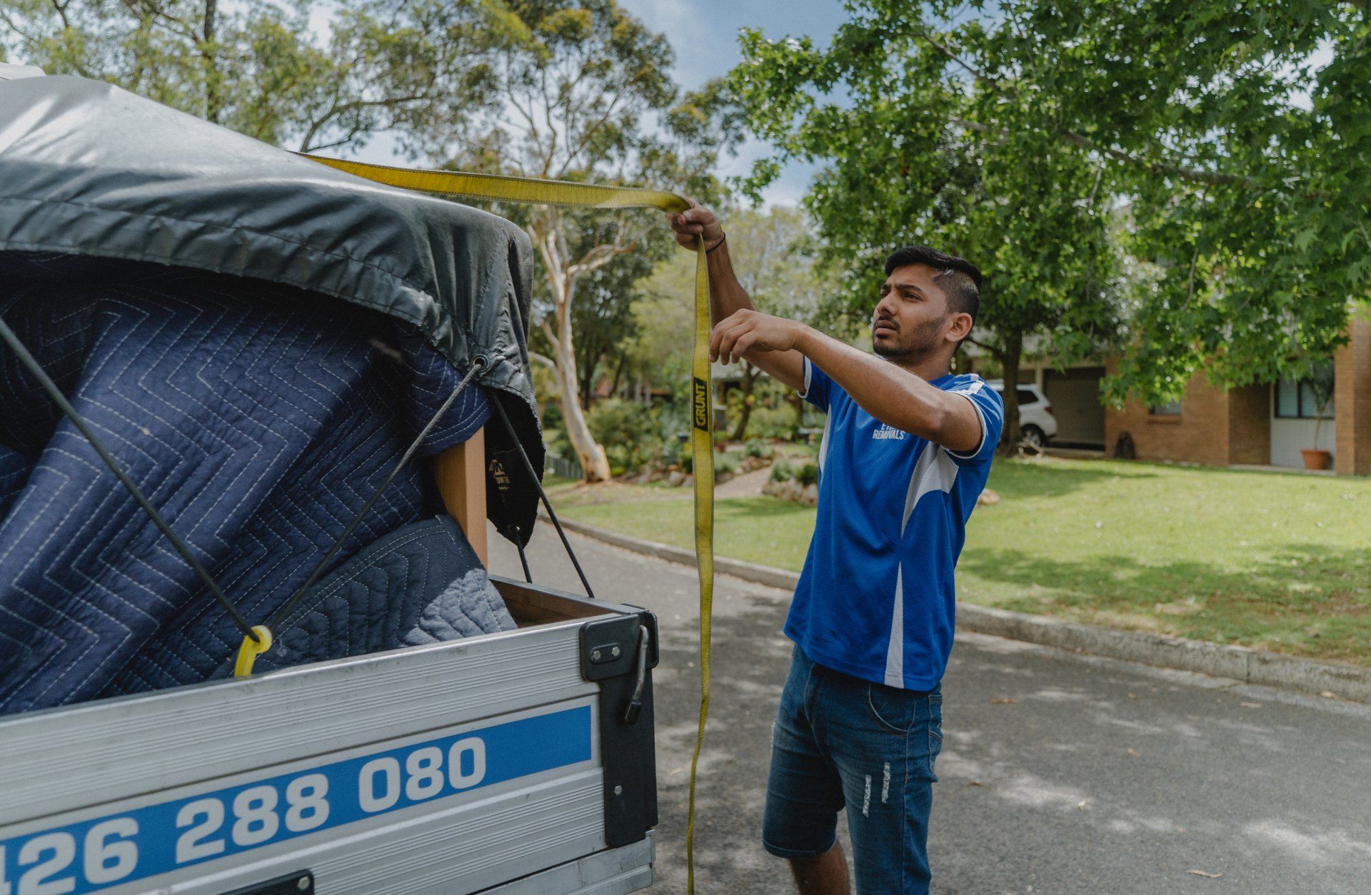 Man in a blue shirt securing a tarp on a loaded truck beside a road under trees