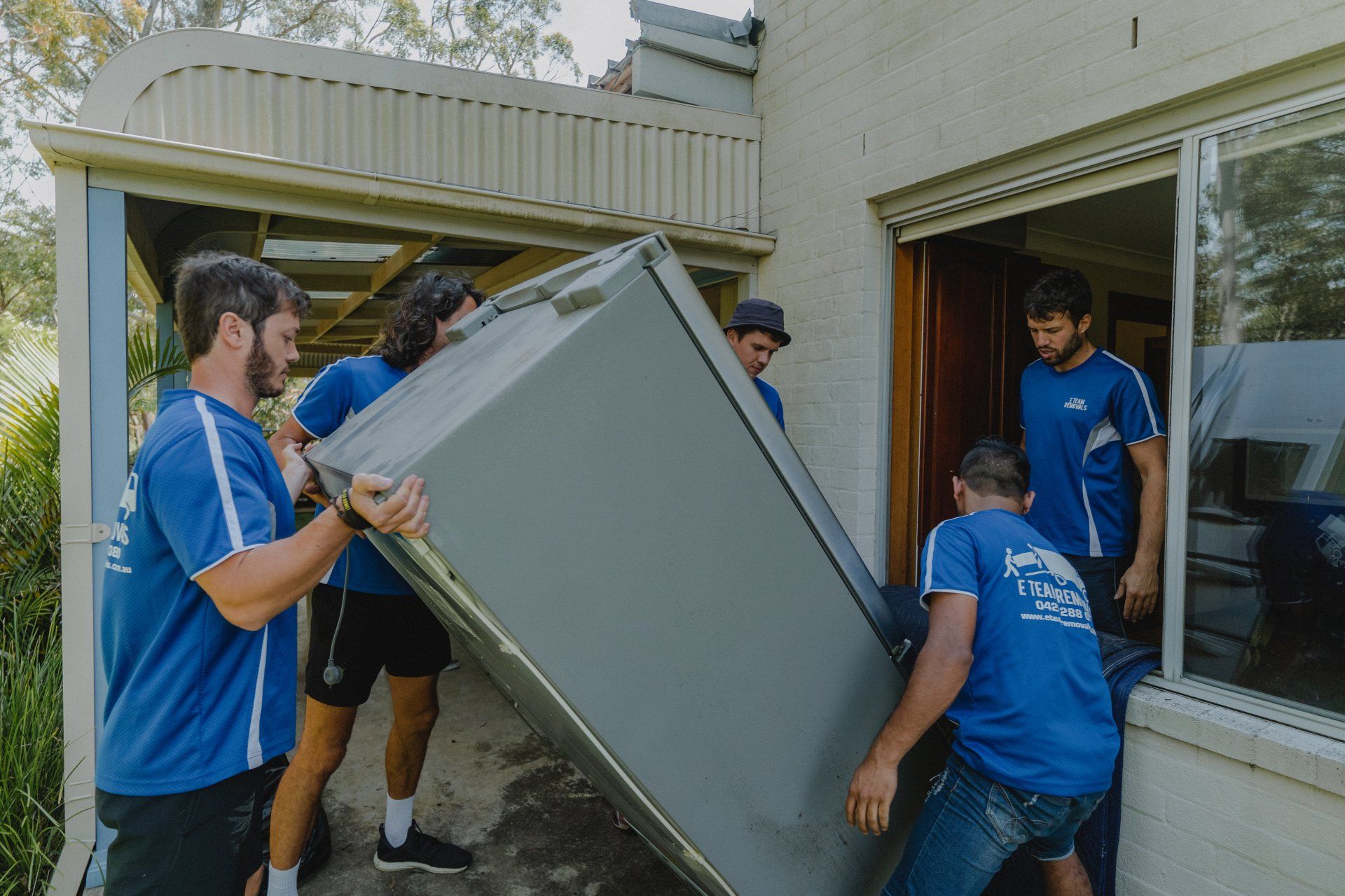 Several people in blue shirts carry a large appliance through a patio door outside a building