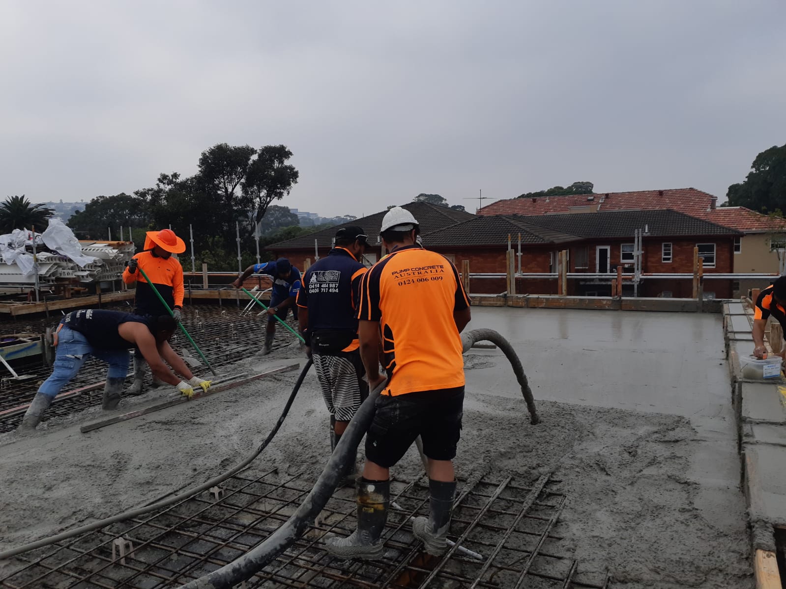 Workers pouring concrete on a construction site beside a brick building, wearing orange safety gear.