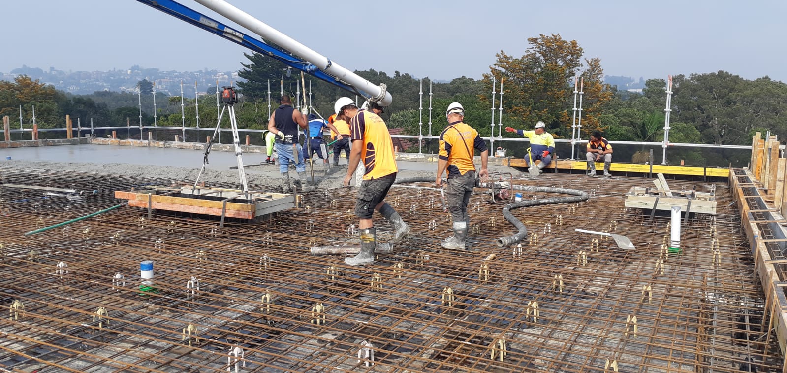 Construction workers in yellow gear on a rebar foundation at a building site, with a crane overhead.