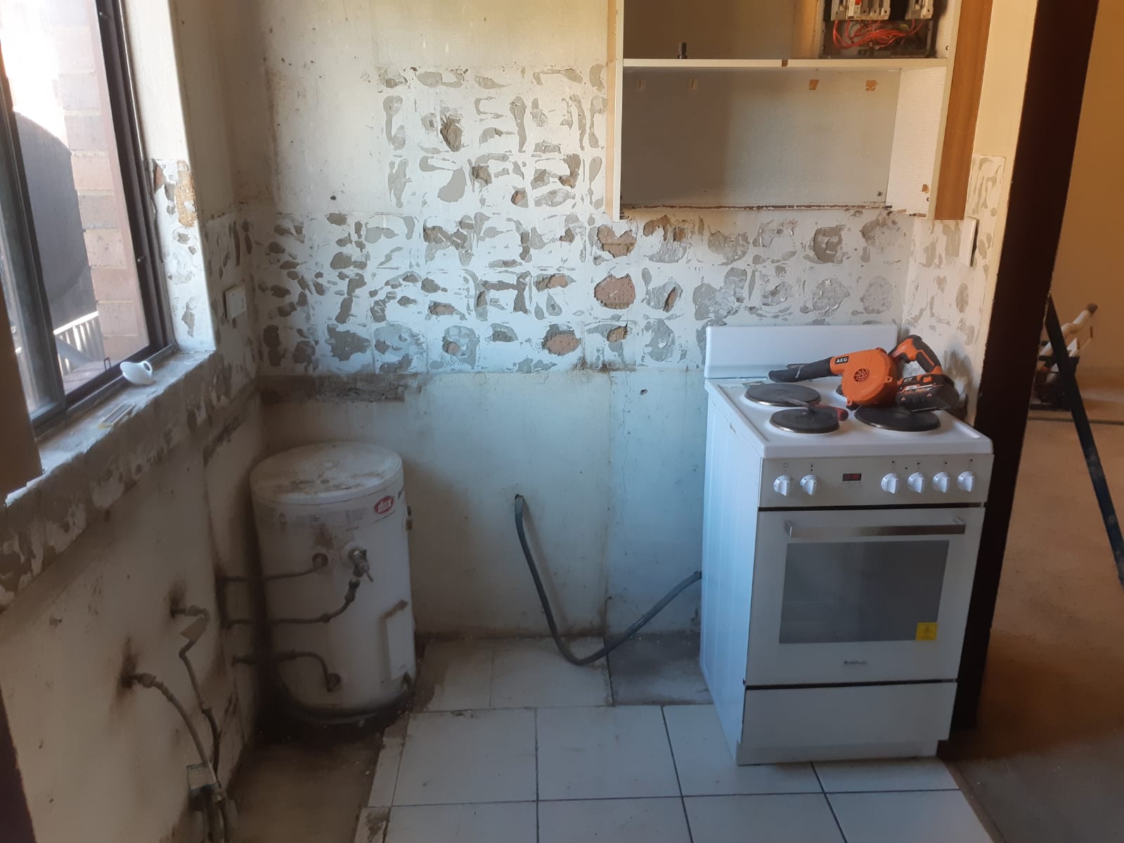 Small kitchen with a stove, water heater, peeling walls, and tiled floor.