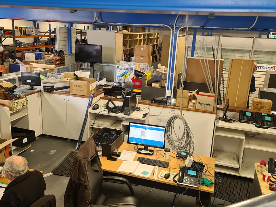 Cluttered office workspace with desks, computer monitors, boxes, and equipment in a busy lab-like room