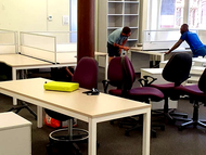 Office workspace with two people working at desks among maroon chairs and cubicles