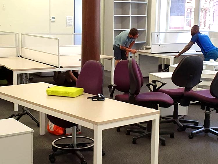 Two people moving office desks and chairs in a workspace with cubicles and purple chairs