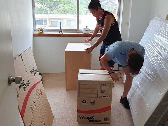 Two people moving boxes in a bright room, with wrapped furniture and a window in the background.