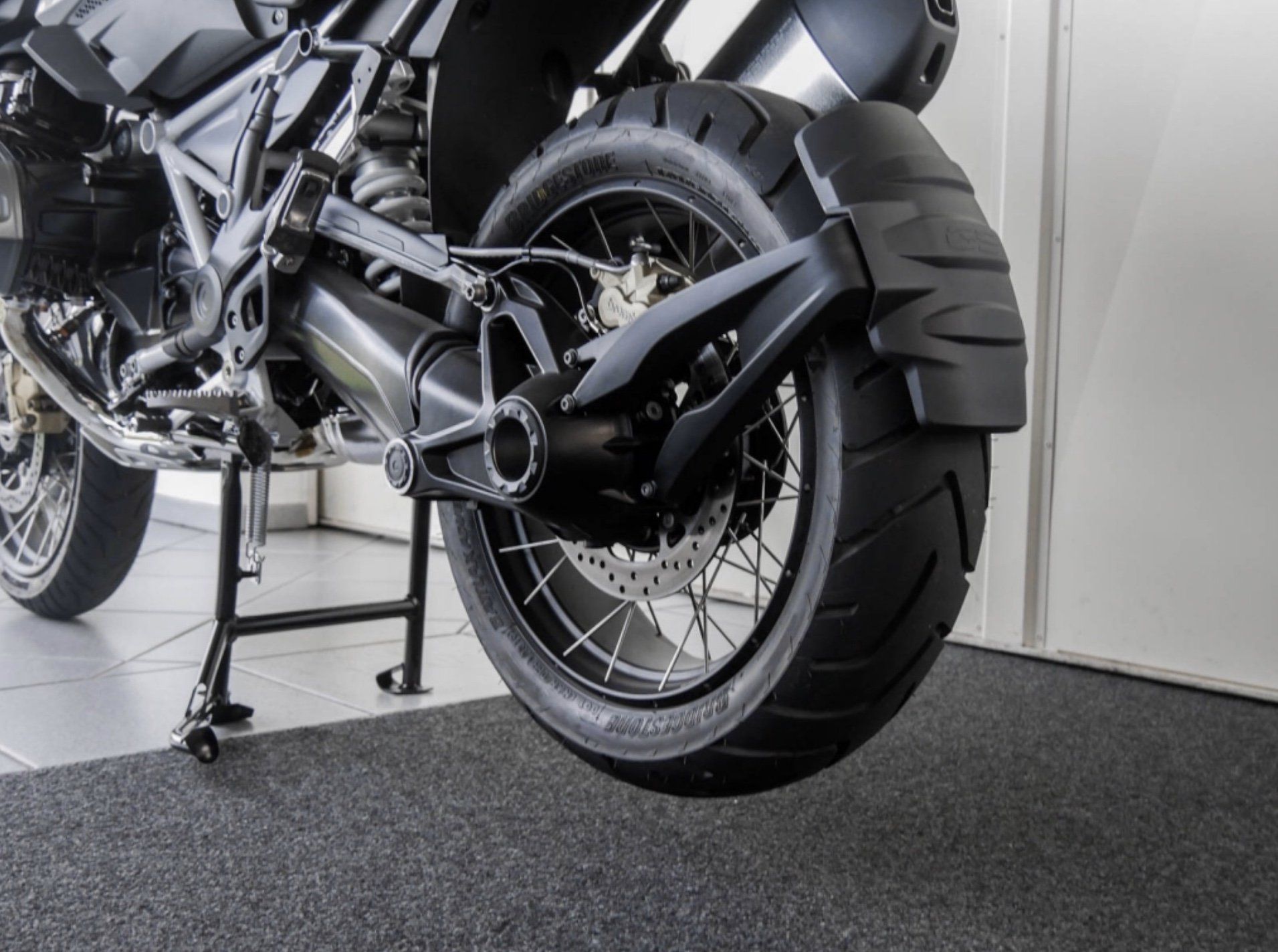Ein Motorrad steht auf Bodenfliesen in der Sicht auf die hintere  Felge vor einer weißen Wand auf dem Ständer.