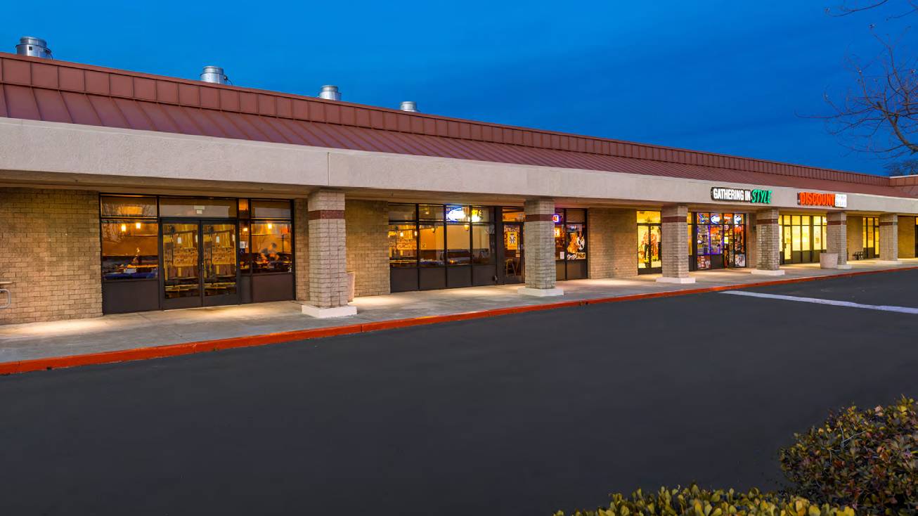 Strip mall storefronts at dusk with lit windows and a mostly empty parking lot