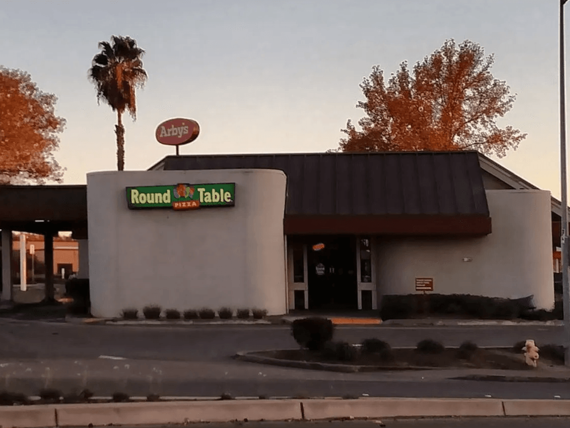 Round Table Pizza storefront at dusk with lit sign and palm trees beside a parking lot