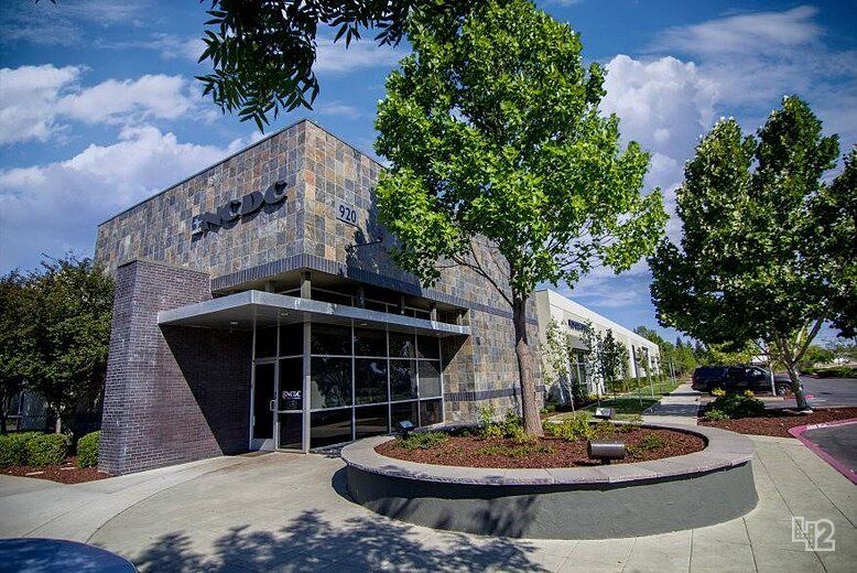 Modern stone office building with glass entrance, trees, and landscaped driveway under blue sky