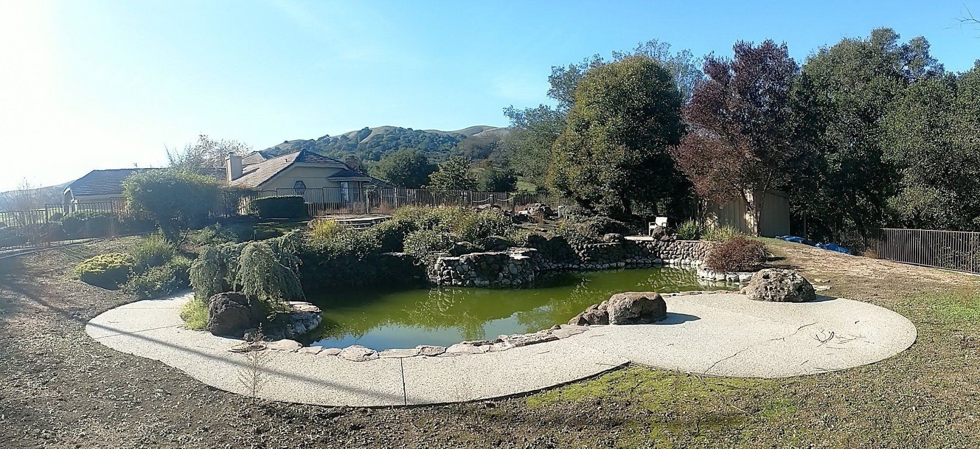 Curved stone path around a small green pond in a sunny landscaped garden with trees and hills