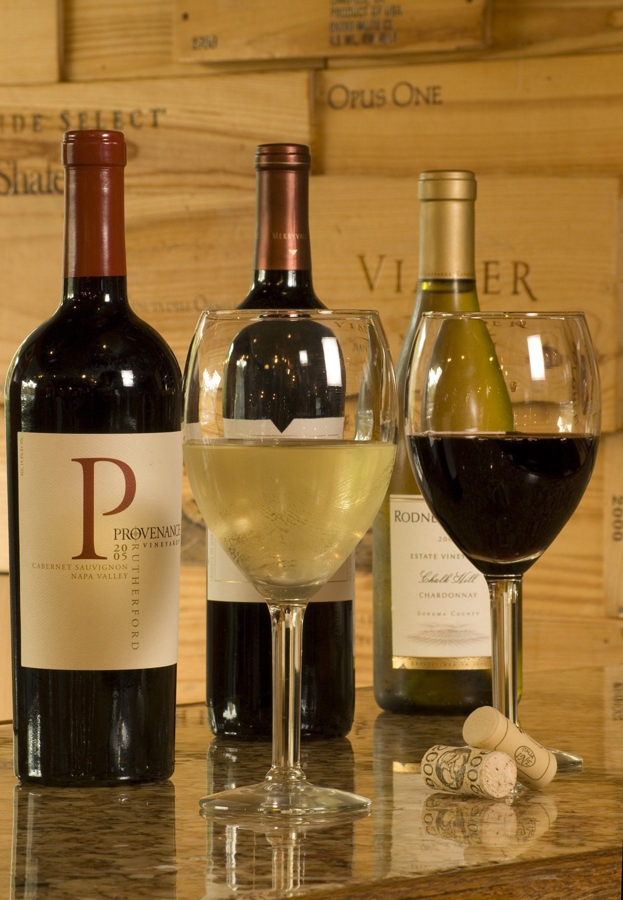 Bottles of red and white wine with two filled glasses, corks in front, wooden backdrop.