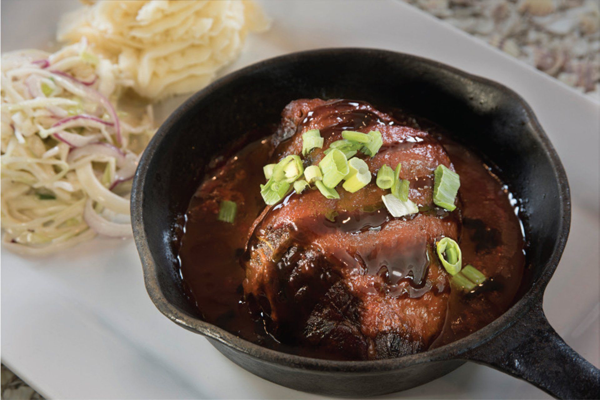 Beef in dark sauce, served in a cast iron skillet, with slaw and mashed potatoes on a white plate.