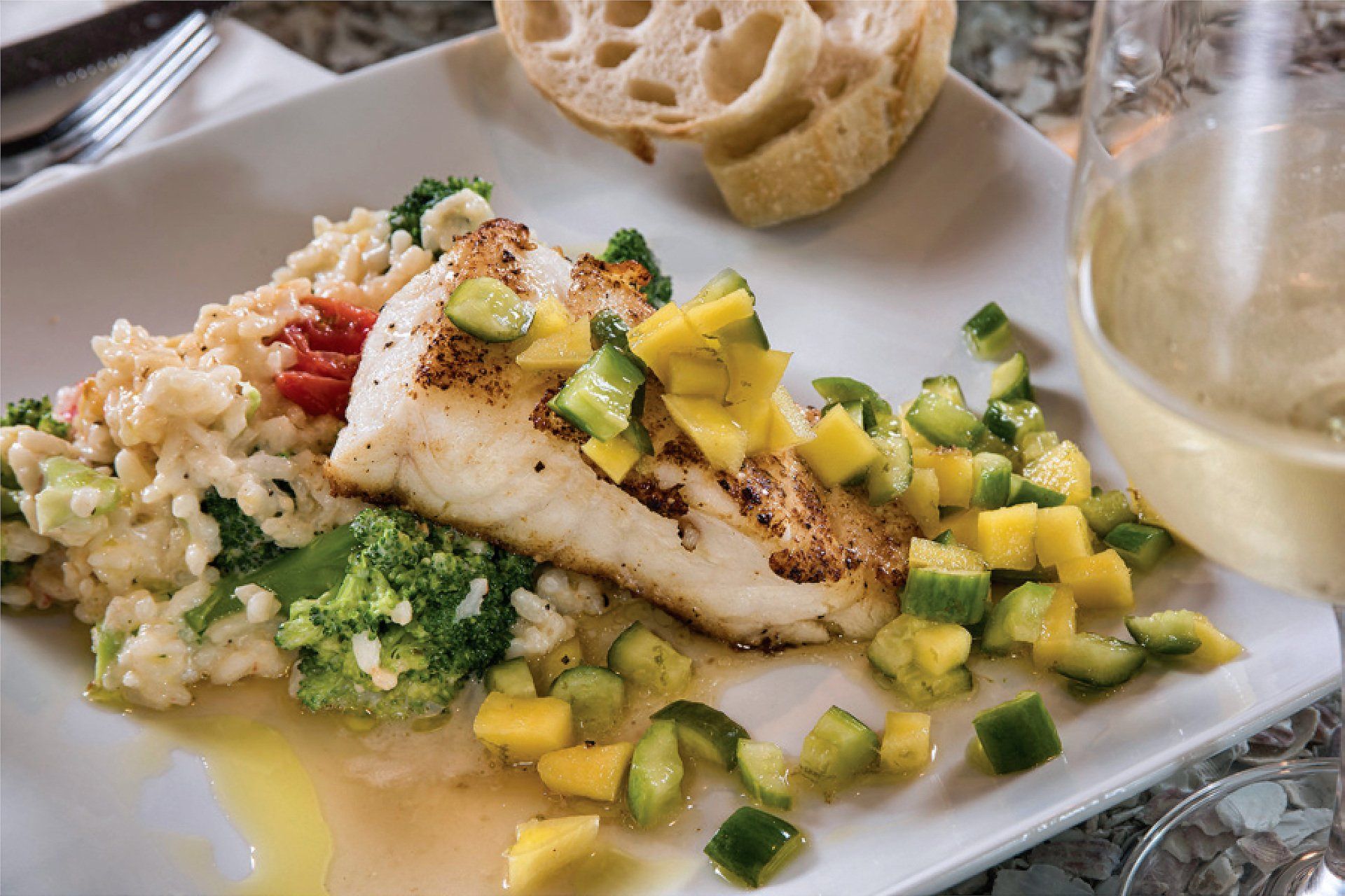 Plate of pan-seared fish with mango salsa, broccoli risotto, and a glass of white wine.