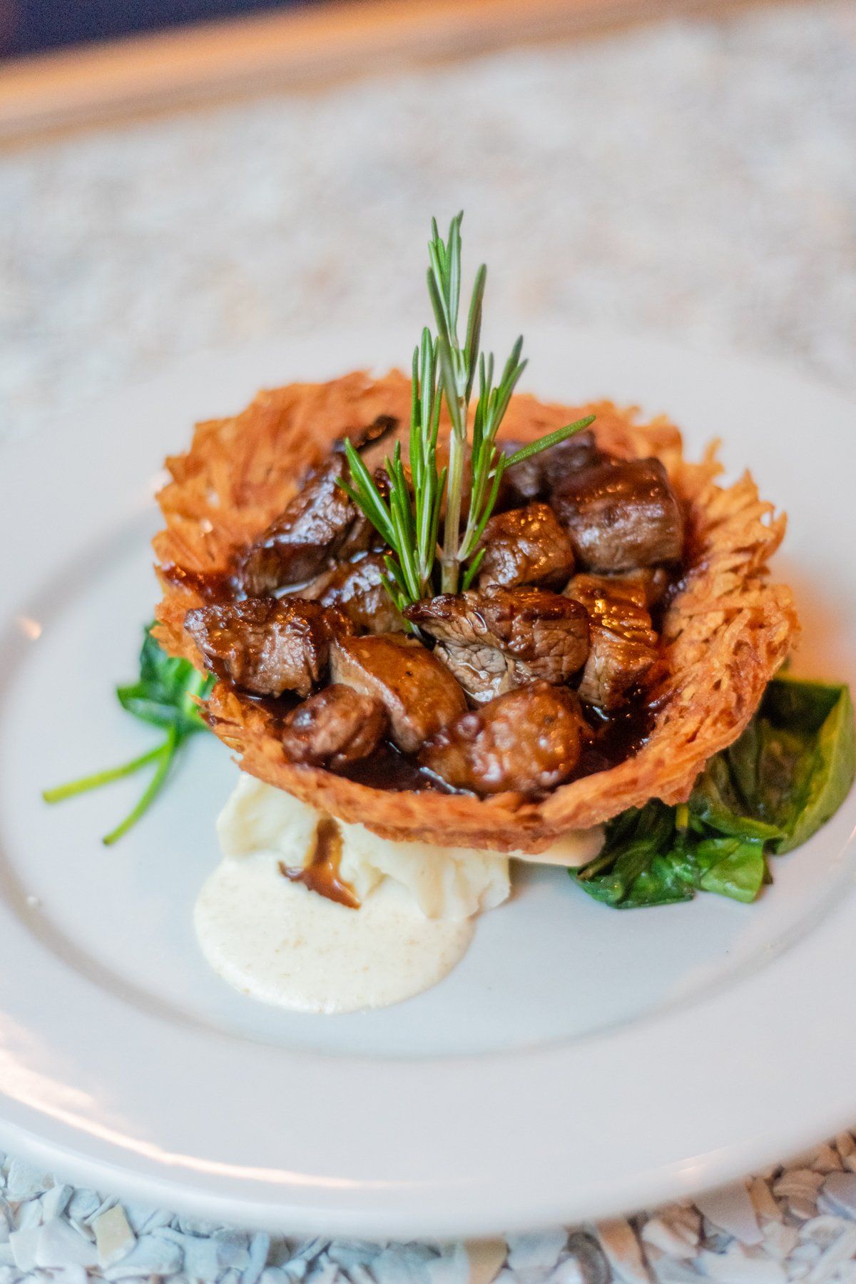 Beef stew in potato basket with rosemary sprig, on mashed potato and spinach, white plate.