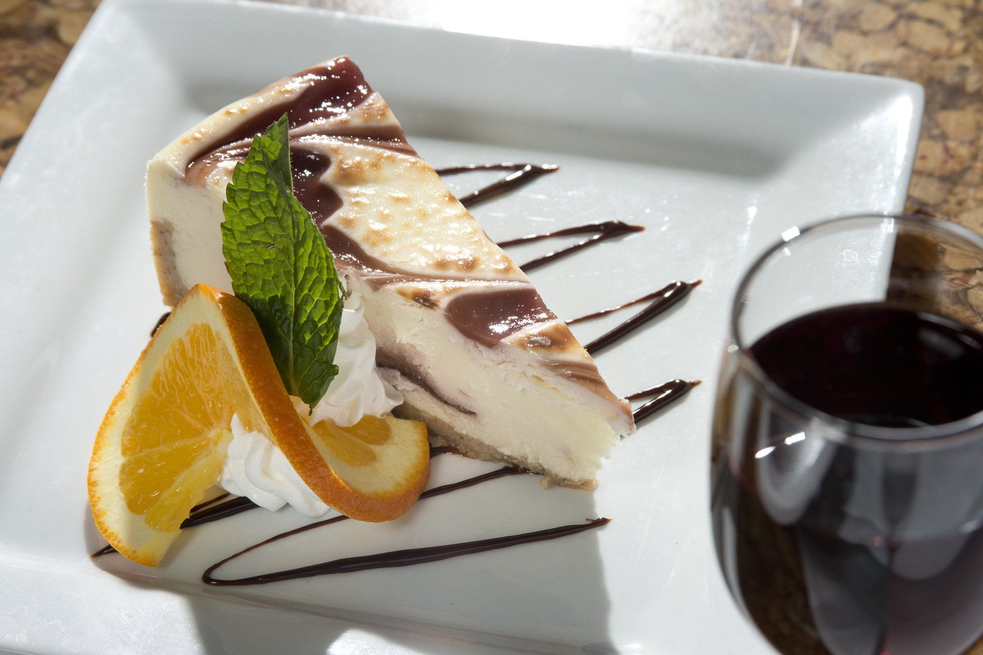 Slice of swirled cheesecake with orange slice, mint, and chocolate drizzle on a white plate, next to a glass of red wine.