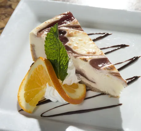 Slice of swirled cheesecake with orange slices, mint, and chocolate drizzle on a white plate.