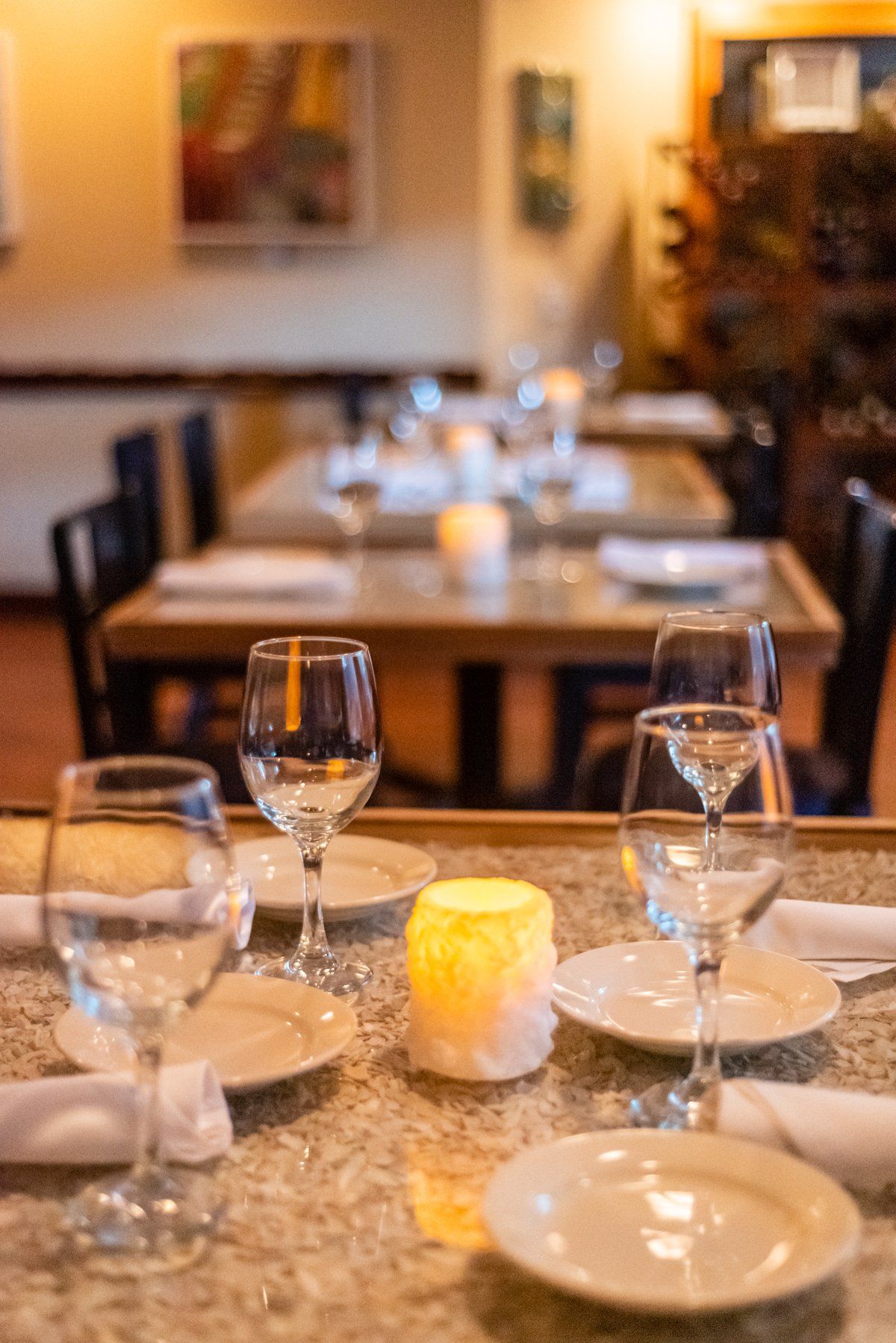 Restaurant table set for dining, with glasses, plates, and a candle, another table in the background.