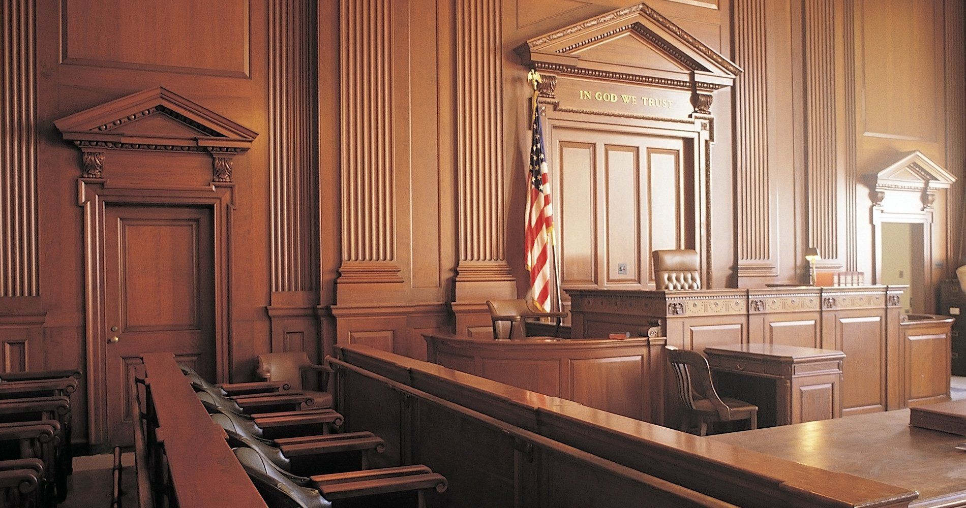 Empty courtroom with wooden benches, judge’s bench, and American flag under warm light.