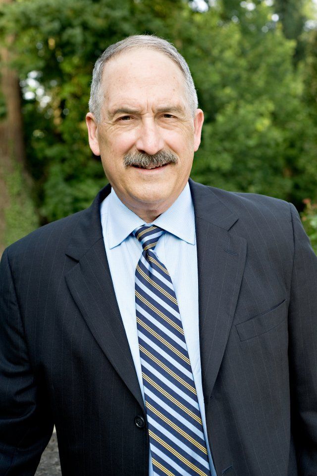 Man in a dark suit and striped tie smiling outdoors with green foliage background