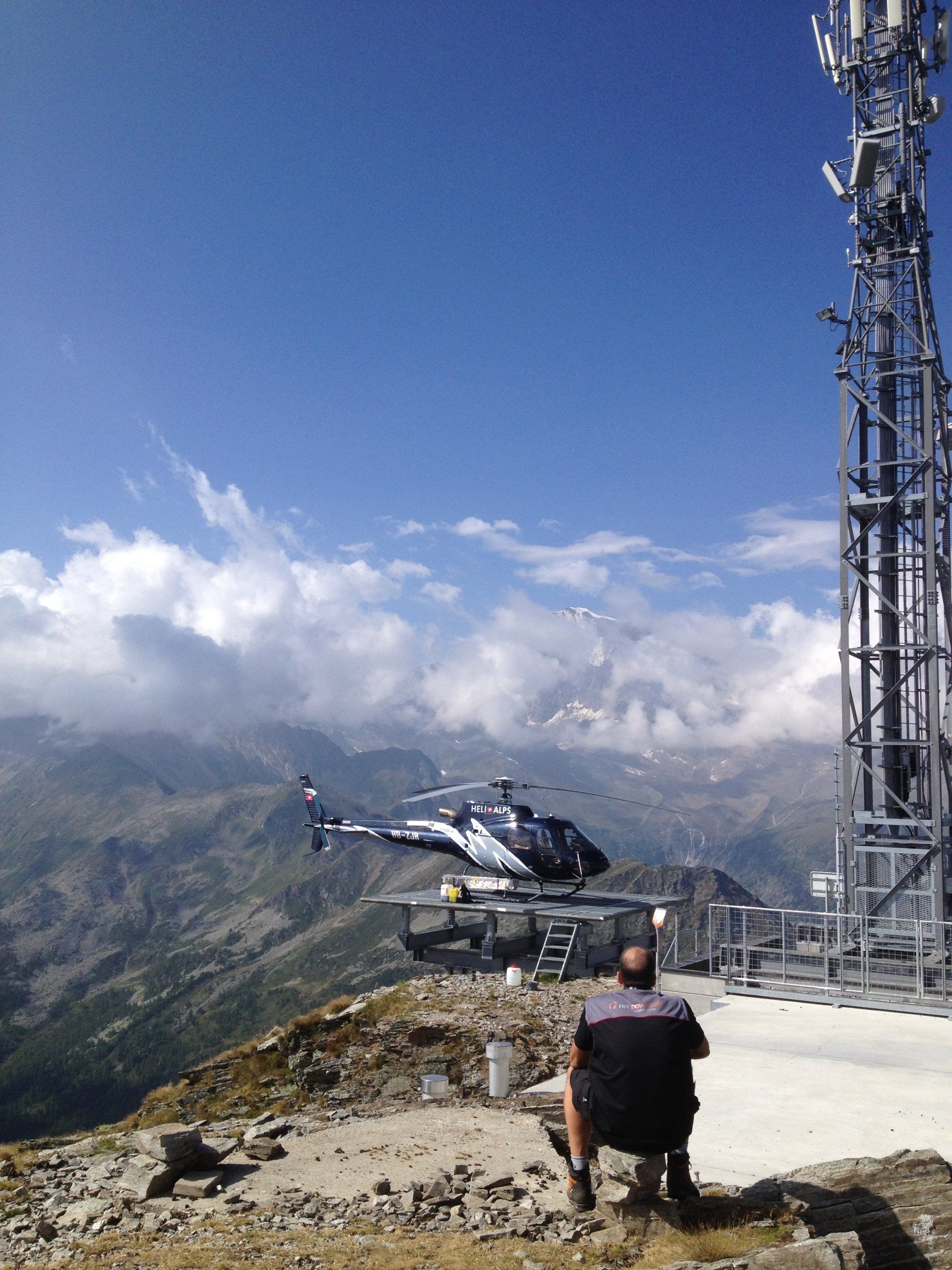 Ein Mann sitzt auf einem Berggipfel und beobachtet einen Hubschrauber beim Abheben