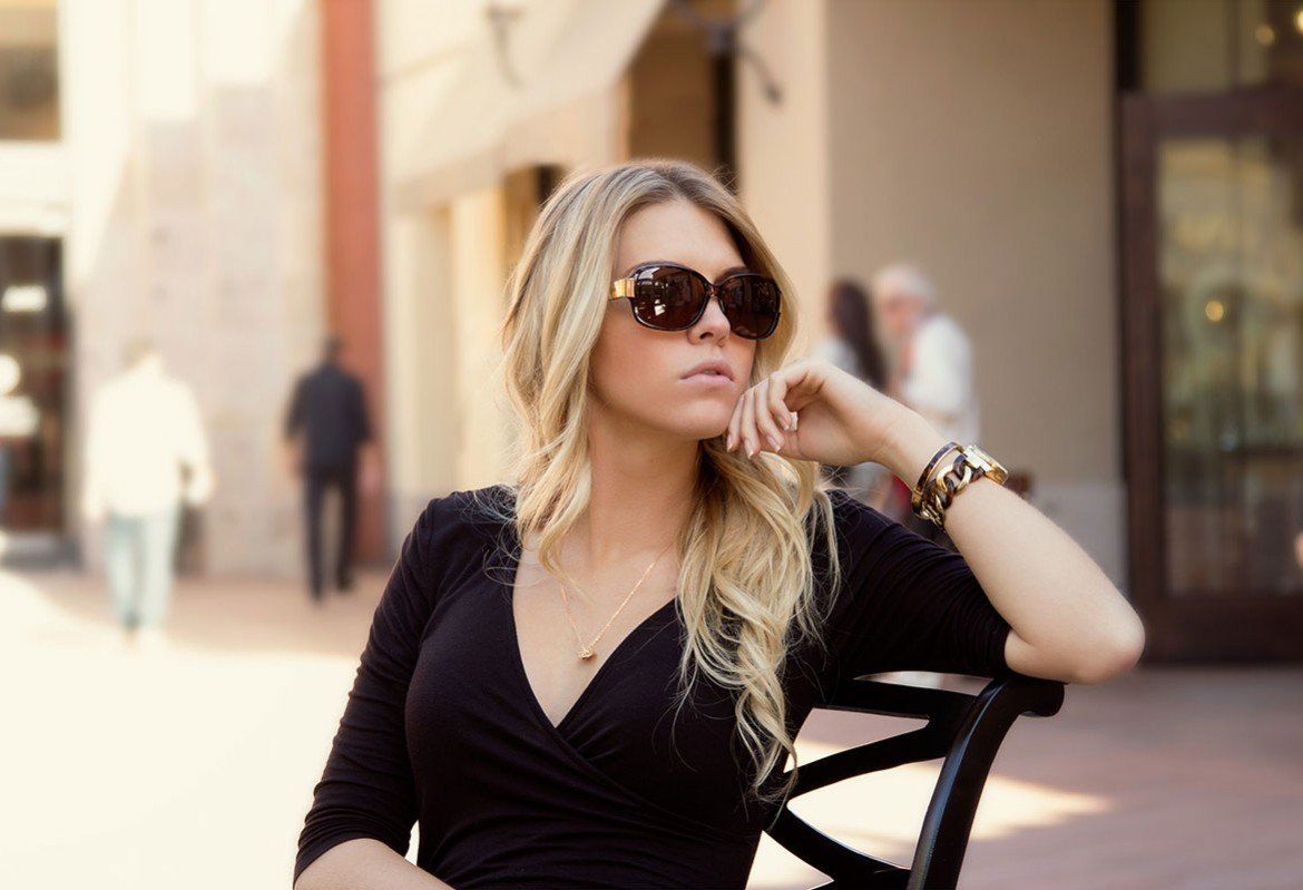 Woman with sunglasses sits outside, resting hand on chin.